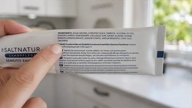 Hand wijst naar de ingrediëntenlijst van een tandpastatube om microplastics te herkennen